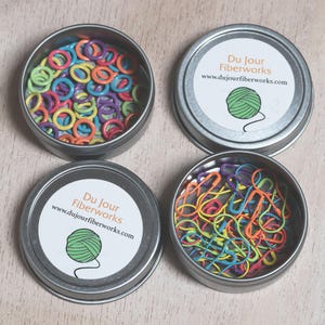 May include: Four round metal tins with lids, each containing colorful plastic stitch markers. The lids have a white label with the text "Du Jour Fiberworks" and a website address.