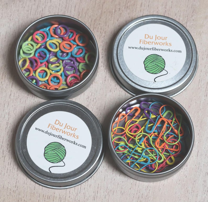 May include: Four round metal tins with lids, each containing colorful plastic stitch markers. The lids have a white label with the text "Du Jour Fiberworks" and a website address.