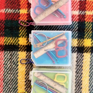 May include: Three clear plastic containers with colorful lids, each containing a set of small tools. The containers are on a red and black plaid surface. The tools include scissors, rulers, crochet hooks, and needles.
