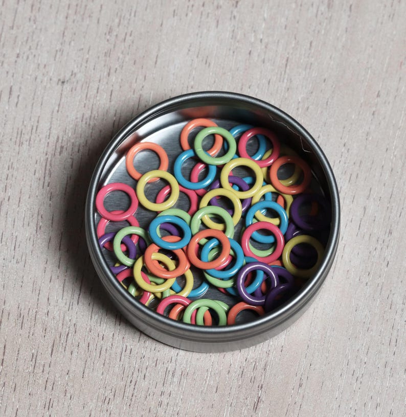 May include: A metal tin filled with colorful plastic rings. The rings are in various shades of pink, orange, yellow, green, blue, and purple.