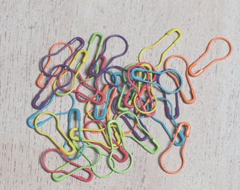 May include: A pile of colorful safety pins in various shades of green, blue, purple, orange, and yellow. The pins are arranged in a random pattern on a light wood surface.