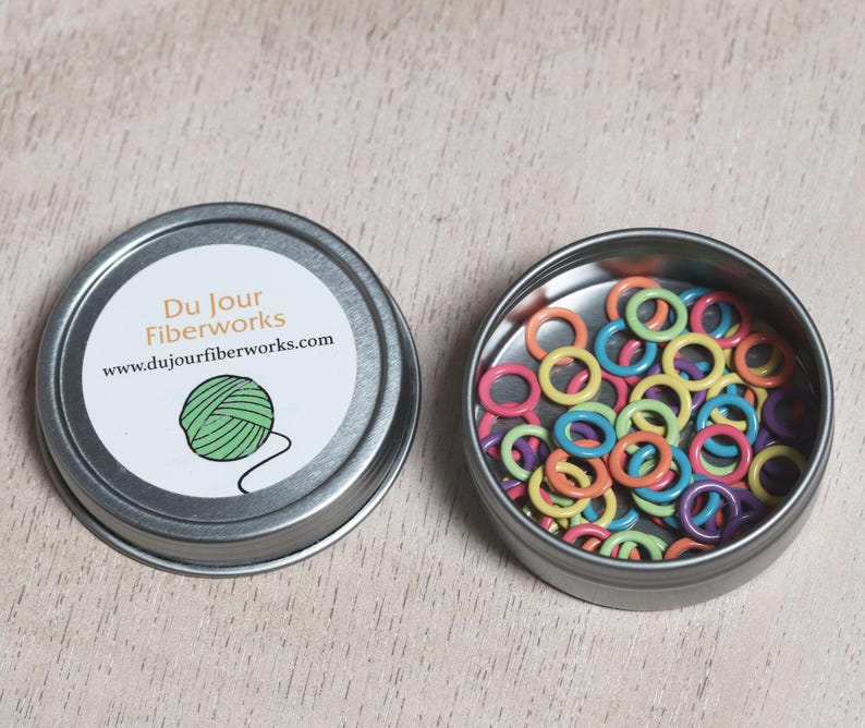 May include: A small tin container with a label that reads "Du Jour Fiberworks" and a website address. The tin is open and contains a variety of colorful plastic stitch markers.