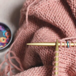 May include: Close-up of a person knitting with gold knitting needles. The yarn is a soft pink color and the needles are resting on a pink knitted fabric.
