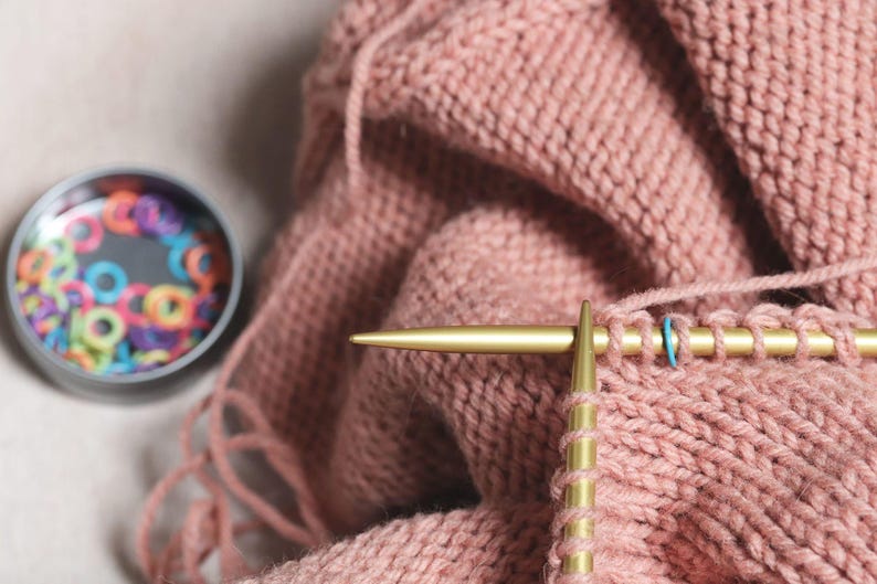 May include: Close-up of a person knitting with gold knitting needles. The yarn is a soft pink color and the needles are resting on a pink knitted fabric.