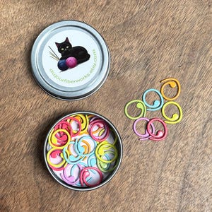 May include: A silver tin with a black cat and yarn ball design, filled with colorful stitch markers. The tin has the text "dujourfiberworks.etsy.com".