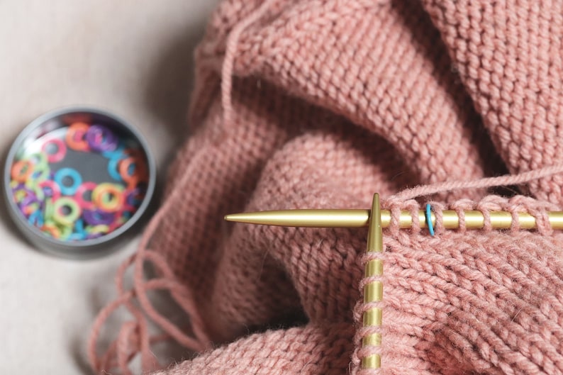 May include: Close-up of a pink knitted fabric with a gold knitting needle and a blue stitch marker. The needle is threaded with yarn and the fabric is being worked on.