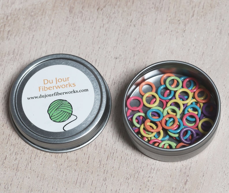 May include: A small tin container with a label that reads "Du Jour Fiberworks" and a website address. The tin is open and filled with colorful plastic stitch markers.