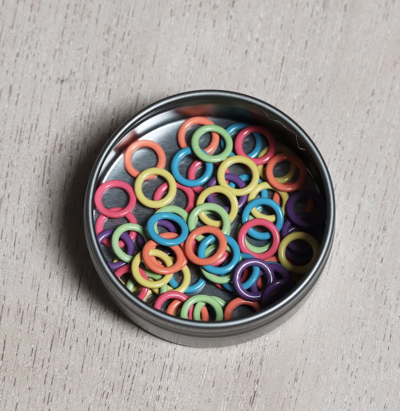 May include: A metal tin filled with colorful plastic rings. The rings are in various shades of pink, orange, yellow, green, blue, and purple.