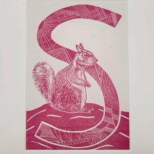 May include: A pink and white linocut print of a squirrel and the letter "S". The squirrel is standing upright, and the letter "S" is behind it. The print is on white paper and framed.
