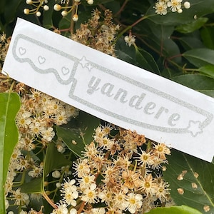 May include: A silver glitter sticker with a knife shape and the word 'yandere' written on it. The sticker has three small hearts and two stars on it.