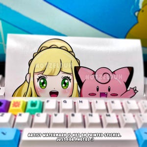 May include: A white sticker featuring two anime-style characters. One is a blonde girl with green eyes, and the other is a pink creature with black ears. The sticker is placed on a white keyboard with colorful keys.