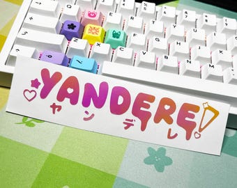 May include: A white mechanical keyboard with custom keycaps in various colors. A sticker with the word "YANDERE" in gradient pink and orange, with Japanese characters, is placed in front of the keyboard.
