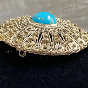 May include: Silver-toned filigree brooch with a central turquoise-colored stone. The brooch has a detailed, openwork design with floral and beaded accents. The pin is oval-shaped and appears to be vintage or antique.