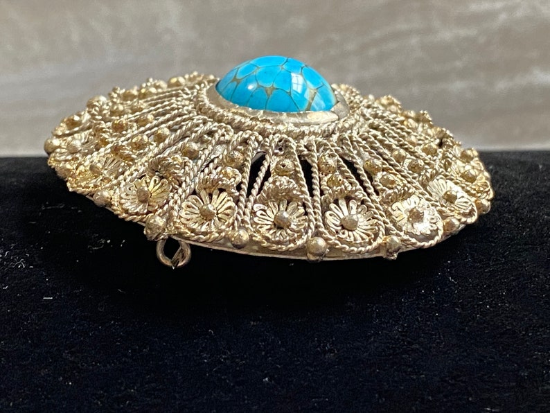 May include: Silver-toned filigree brooch with a central turquoise-colored stone. The brooch has a detailed, openwork design with floral and beaded accents. The pin is oval-shaped and appears to be vintage or antique.