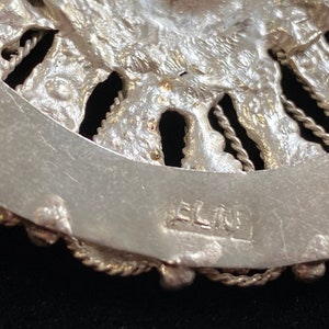 May include: Close-up of a silver-toned, ornate jewelry piece. The design features a detailed, textured pattern with a circular shape. The piece has a decorative border and the word "ELIN" is stamped on the back.