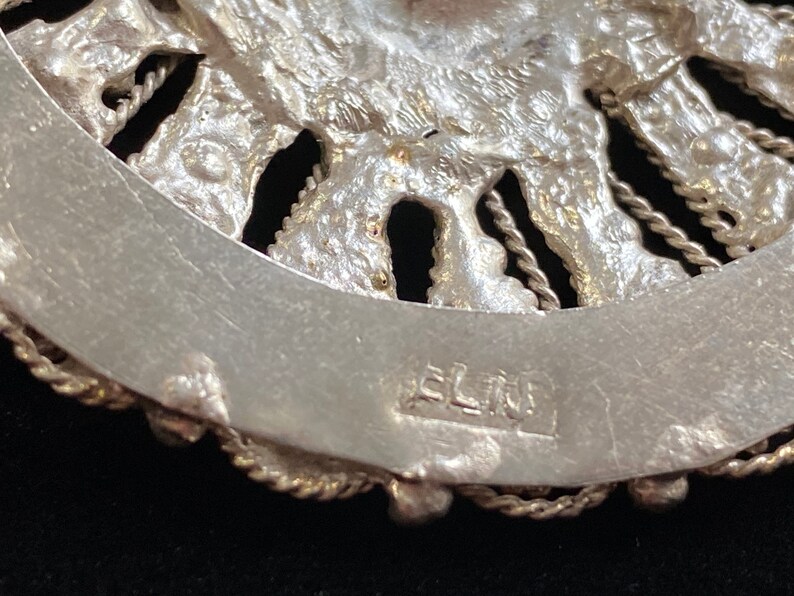 May include: Close-up of a silver-toned, ornate jewelry piece. The design features a detailed, textured pattern with a circular shape. The piece has a decorative border and the word "ELIN" is stamped on the back.
