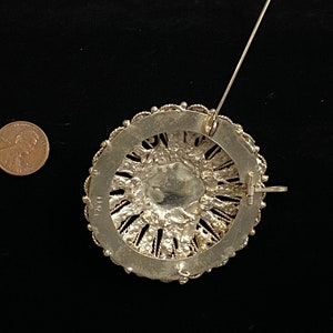 May include: A silver-toned brooch with a circular design. The brooch features intricate filigree work around a central, possibly glass, element. A penny is included for size reference. The pin and clasp are visible on the back.