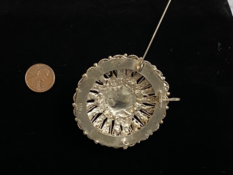 May include: A silver-toned brooch with a circular design. The brooch features intricate filigree work around a central, possibly glass, element. A penny is included for size reference. The pin and clasp are visible on the back.