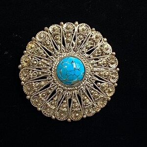 May include: A detailed, silver-toned brooch with a central, round turquoise stone. The brooch features an intricate, openwork design with floral and beaded elements. The turquoise stone is a vibrant blue, set against the metallic silver. The brooch is circular in shape.