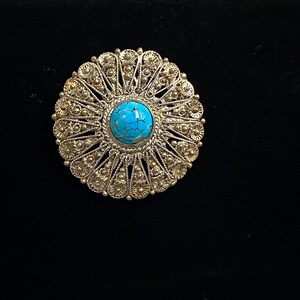 May include: A silver-toned, circular brooch with a bright turquoise center stone. The brooch features an intricate, openwork design with a floral pattern around the edges. The jewelry is set against a black background.