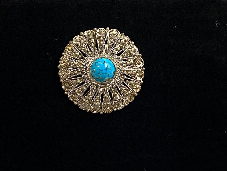 May include: A silver-toned, circular brooch with a bright turquoise center stone. The brooch features an intricate, openwork design with a floral pattern around the edges. The jewelry is set against a black background.