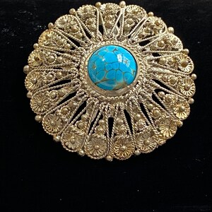 May include: A silver-toned brooch with a central, round, turquoise-colored stone. The brooch features an intricate, openwork design with floral and beaded details. The background is black.