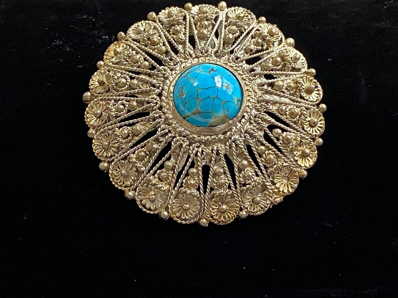 May include: A silver-toned brooch with a central, round, turquoise-colored stone. The brooch features an intricate, openwork design with floral and beaded details. The background is black.