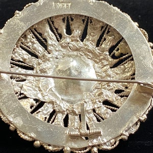 May include: A detailed view of the back of a silver brooch. The brooch has a circular design with a central, textured element surrounded by a decorative border. The pin and clasp are visible. The brooch has a vintage aesthetic.