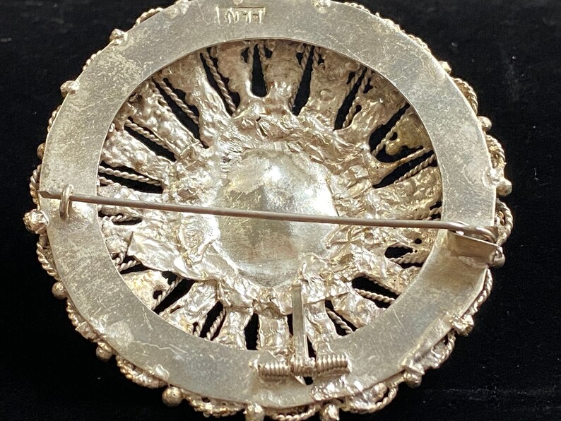 May include: A detailed view of the back of a silver brooch. The brooch has a circular design with a central, textured element surrounded by a decorative border. The pin and clasp are visible. The brooch has a vintage aesthetic.