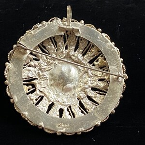 May include: A silver-toned brooch with a circular design. The brooch features a central, textured element surrounded by a detailed, openwork border. A pin and a loop for a chain are visible on the back. The brooch has a vintage aesthetic.