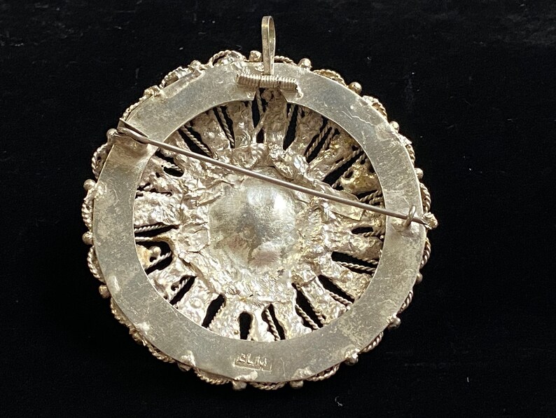 May include: A silver-toned brooch with a circular design. The brooch features a central, textured element surrounded by a detailed, openwork border. A pin and a loop for a chain are visible on the back. The brooch has a vintage aesthetic.
