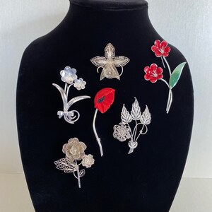May include: A collection of floral brooches displayed on a black mannequin. The brooches feature various designs, including a red poppy, silver filigree flowers, and red enamel flowers. The brooches are made of metal and are suitable for adding a touch of elegance to any outfit.