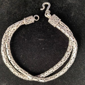 May include: A silver bracelet with a three-strand Byzantine chain design. The bracelet features an ornate hook clasp and decorative end pieces. The jewelry is displayed against a dark background, highlighting its intricate details.