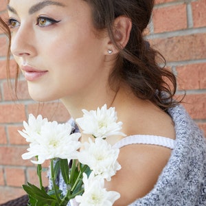 May include: A close-up of a woman's face with white flowers in front of her. She is wearing a gray sweater and has a small heart-shaped earring in her left ear.