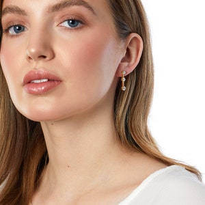 Könnte beinhalten: Goldfarbene Ohrhänger mit drei klaren, funkelnden Edelsteinen. Die Ohrringe sind mit einer linearen Anordnung der Edelsteine gestaltet, wodurch ein zarter und eleganter Look entsteht. Die Ohrringe werden von einer Person getragen.