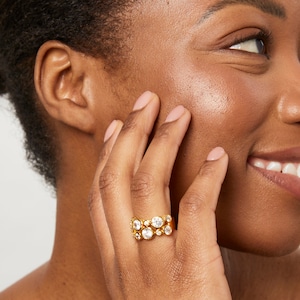 May include: A gold-toned ring featuring a wide band adorned with multiple round, clear, faceted stones of varying sizes arranged in a cluster pattern across the surface, creating a textured and reflective appearance on the finger.