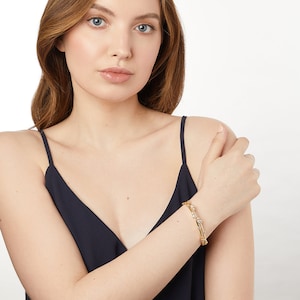 Könnte beinhalten: Ein goldenes Armreif-Armband mit kleinen, klaren Steinen, die in das Metall gefasst sind. Das Armband befindet sich an einem Handgelenk.