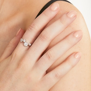 Op de afbeelding: Een zilverkleurige ring met vier ronde, heldere, gefacetteerde stenen van verschillende groottes, gerangschikt in een cluster langs de band. De stenen zijn gezet in gladde, cirkelvormige metalen zettingen voor een asymmetrisch ontwerp.