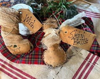 Op de afbeelding: Drie handgemaakte kerstversieringen met jute stof en bruine papieren etiketten. De etiketten zijn gelabeld met "Wish", "Sleigh Bells" en een derde etiket met onleesbare tekst. De versieringen liggen op een rood-zwarte geruite stof en een witte stof met rode strepen.