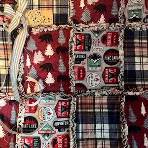 May include: A rag quilt featuring square patches with patterns of black bears, pine trees, plaid fabric, and outdoor adventure badges. The badges include text such as Bear Creek, Pine Lake, and Basecamp. A light-colored striped ribbon is attached.