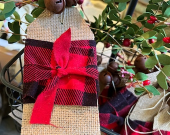 May include: A set of burlap gift tags with red and black buffalo plaid ribbon bows. The tags are decorated with small, rustic bells and are displayed in a wire basket.