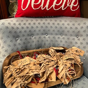 Op de afbeelding: Een houten kom gevuld met handgemaakte kerstversiering. De versiering is gemaakt van bruine stofstroken en kaneelstokjes. Elke versiering heeft een handgeschreven label met het woord "Vrede" erop. De kom staat op een blauwe fauteuil met een rood kussen waarop in witte letters "geloven" staat.