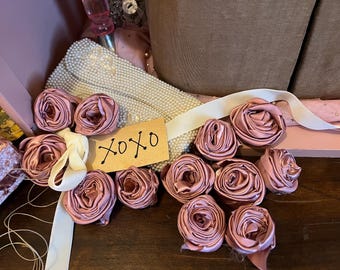 May include: A collection of dusty rose fabric roses with a white ribbon and a brown card with the text "XOXO" on a wooden surface.