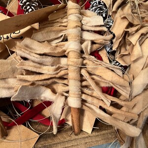 Op de afbeelding: Een kaneelstokje gewikkeld in beige en witte stofstroken, omringd door bruine papieren etiketten, rood lint en dennenappels.
