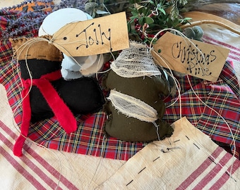 May include: Three handmade Christmas ornaments on a plaid fabric background. The ornaments are a black boot with a red ribbon, a green tree with a white gauze overlay, and a brown tree with a white gauze overlay. The ornaments have brown paper tags with the words "Jolly", "Christmas", and "Pine" written on them.