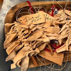 Op de afbeelding: Een houten kom gevuld met handgemaakte stoffen ornamenten. De ornamenten zijn gemaakt van beige stofstroken en hebben kaartjes met de tekst "Peace" erop.