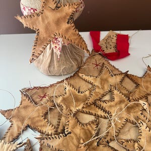 May include: A collection of rustic, handmade star ornaments. The stars are crafted from tan-colored fabric with black stitching and a distressed appearance. Some stars are attached to a burlap-wrapped base, and others are strung together with twine. A red tag with the word "shine" is also present.