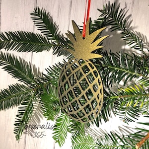 May include: Gold glitter pineapple Christmas bauble hanging from a red ribbon against a white wood background with green pine branches.