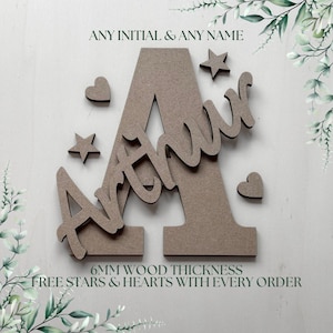 May include: A large wooden letter A with the name Arthur in cursive script layered on top. The design includes small wooden stars and hearts. The text ANY INITIAL & ANY NAME and 6MM WOOD THICKNESS are visible. The material has a matte brown finish.