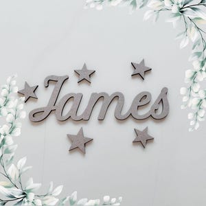 May include: Wooden name sign with the name "James" in a cursive font, accompanied by several star-shaped cutouts. The sign and stars are a muted gray color, set against a light gray background with decorative floral accents.